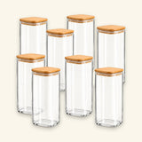 Six clear glass canisters with square bases and light natural wood lids, arranged together on a plain light beige background.