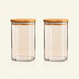 Two clear glass canisters with square bases and light natural wood lids, shown side by side on a plain off-white background.