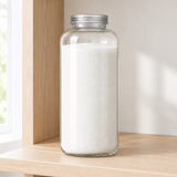 A tall clear glass jar filled with white sugar, sealed with a silver metal lid, standing on a light wood shelf in a bright indoor setting.