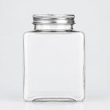 A clear glass jar with a square shape and a silver metal screw-on lid, shown against a plain light gray background.