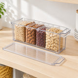 Clear plastic divided storage container filled with assorted nuts and dried fruits, shown on a light wood countertop with its lid placed in front. A plant and stacked dishes are visible in the background.