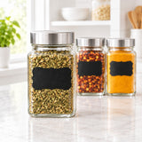Three clear glass spice jars with silver metal lids and black chalkboard-style labels, filled with dried herbs, red pepper flakes, and yellow-orange spices, arranged on a light marble countertop in a bright kitchen setting.