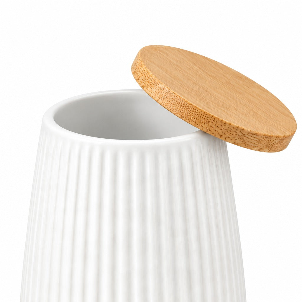 A white ribbed ceramic jar with a round bamboo lid leaning against it on a light gray studio background.