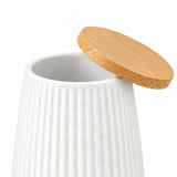 A white ribbed ceramic jar with a round bamboo lid leaning against it on a light gray studio background.