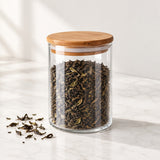 A clear glass tea jar with a light brown wooden lid, filled with loose-leaf tea, set on a white marble surface with a few tea leaves scattered nearby.