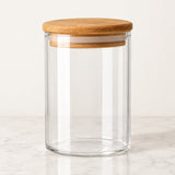 A clear glass canister jar with a round natural bamboo lid, shown on a light marble surface against a neutral off-white background.