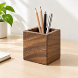 A square wooden pencil holder with a rich dark brown wood grain finish, filled with three pencils and two pens, sitting on a light wood desk next to a potted plant.