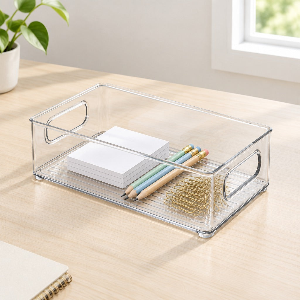 Clear plastic desk organizer tray with side handles, shown on a light wood desk holding white sticky notes, colored pencils, and gold paperclips.