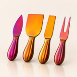 Four metallic cheese knives and a fork with iridescent handles, arranged in a row on a light beige background. The handles transition through pink, orange, and gold tones, and the blades have matching gradient finishes.