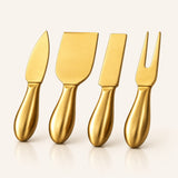 Four metallic gold cheese knives and spreaders with smooth handles, arranged in a row on a clean off-white background.