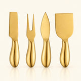 Four gold-colored cheese and charcuterie utensils with matching handles, including a knife, a two-pronged fork, a spreader, and a large scraper, arranged in a row against a light cream background.