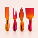 Four iridescent metal cheese and carving tools with handles, arranged in a row on a light beige background. The set includes a knife, a two-pronged fork, a spreader, and a larger spatula, all featuring a vibrant orange, yellow, and pink metallic gradient finish.