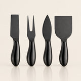 Four black cheese knives with glossy handles, including a flat blade, a two-pronged fork, a spear tip, and a broad spatula, arranged in a row on a light beige background.