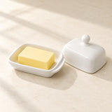 A white ceramic butter dish with a matching lid and knob, shown on a light beige surface with a yellow bar of butter inside the dish. The scene is softly lit with natural shadows.
