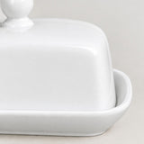 A close-up of a glossy white ceramic butter dish with a matching lid and a small side handle, set against a neutral light beige background.