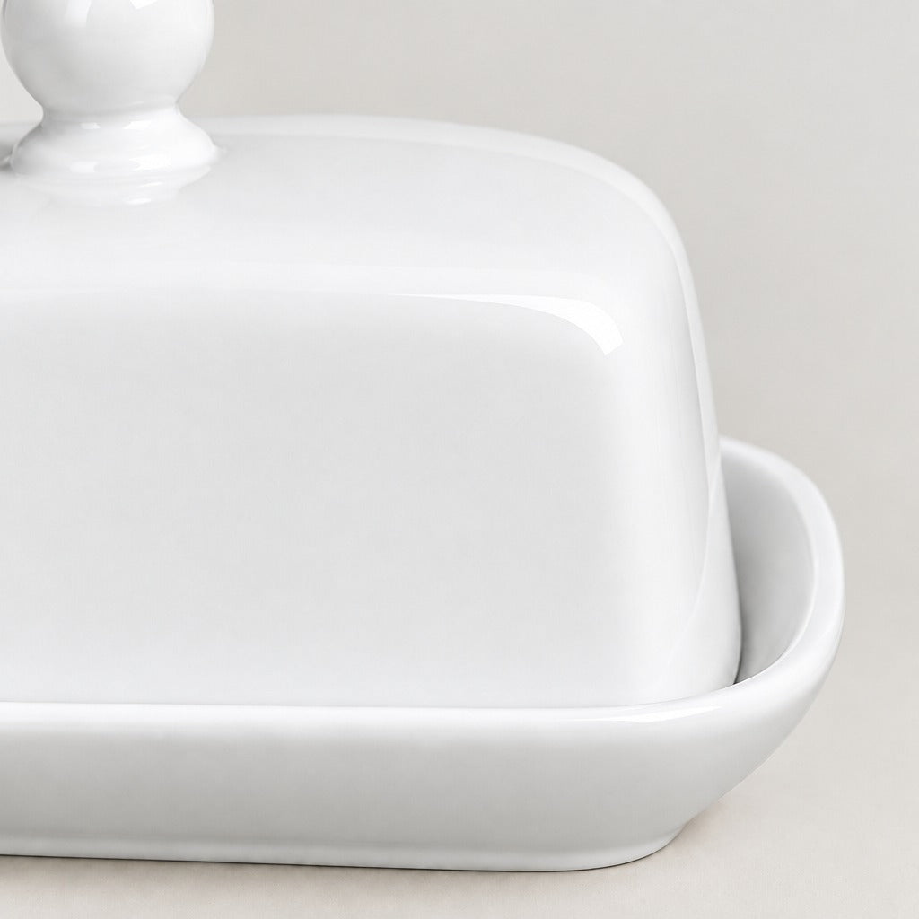 A close-up of a glossy white ceramic butter dish with a matching lid and a small side handle, set against a neutral light beige background.