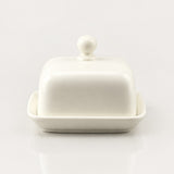 A white ceramic butter dish with a matching lid featuring a round knob and a shallow side dish base, shown on a clean white background with a soft reflection below.