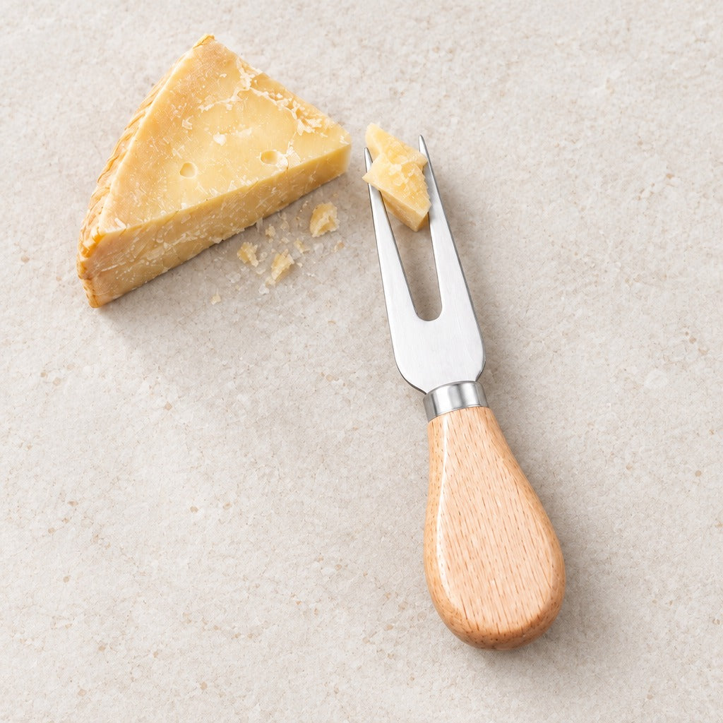 A cheese knife with a light wood handle and stainless steel forked blade, shown with a wedge of hard cheese on a light beige stone surface.