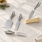 Four-piece stainless steel cheese board utensil set with a spatula, knife, fork, and smaller knife, arranged on a textured beige surface next to a wedge of brie cheese, olives, and rosemary.