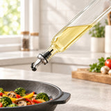 A clear glass oil dispenser bottle with a metal pour spout, filled with golden olive oil, shown dispensing oil over a black skillet containing broccoli, mushrooms, and peppers in a bright kitchen.