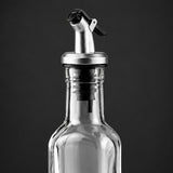 A close-up of a clear glass bottle with a stainless steel pour spout and black flip-top lid, shown against a dark gray background.