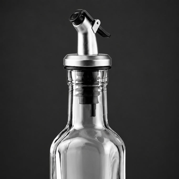 A close-up of a clear glass bottle with a stainless steel pour spout and black flip-top lid, shown against a dark gray background.