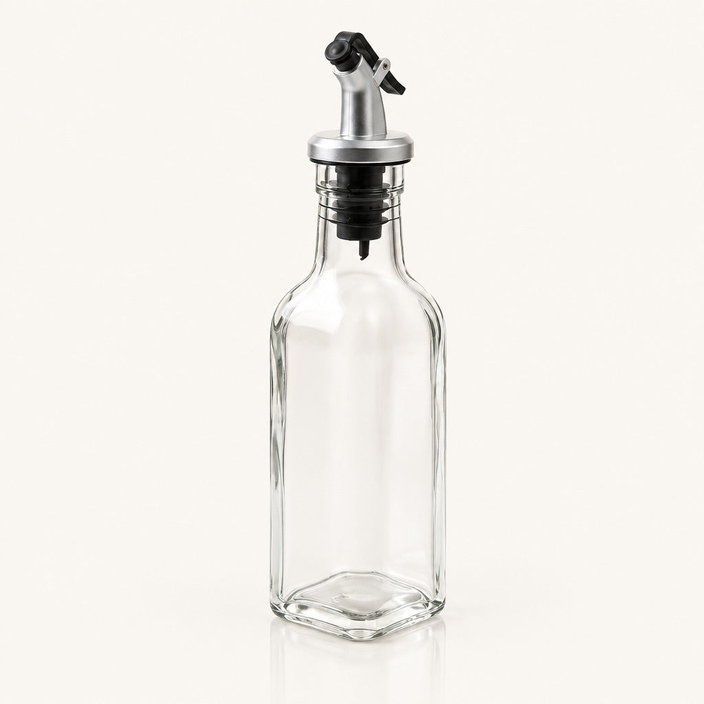 A clear glass bottle with a hexagonal base and a silver metal pour spout with a black plastic cap, shown on a light cream background.