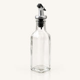 A clear glass bottle with a hexagonal base and a silver metal pour spout with a black plastic cap, shown on a light cream background.