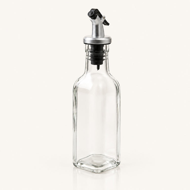 A clear glass bottle with a hexagonal base and a silver metal pour spout with a black plastic cap, shown on a light cream background.