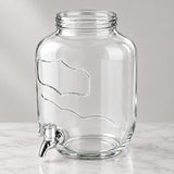 Clear glass beverage dispenser with a wide mouth, embossed wavy patterns, and a chrome metal spout, shown on a light marble surface against a neutral gray background.