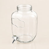 Clear glass beverage dispenser jar with a screw-top neck, side handle cutout, and a small metal spigot, shown on a plain warm beige background.