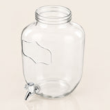 Clear glass beverage dispenser can with a metal spout, photographed on a plain light beige background.