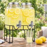 Two large glass lemonade dispensers filled with yellow lemonade and lemon slices, displayed on a black metal stand on a wooden table outdoors with a glass being filled and lemons nearby.