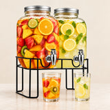 Two large glass beverage dispensers filled with fruit and lemonade on a black metal stand, with two clear glasses of the drink in front, photographed on a light beige background.