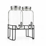 Two clear glass beverage dispensers with silver metal lids and spouts, displayed on a black wire stand with an additional clear measuring cup on a white background.