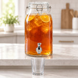 A large clear glass iced tea dispenser with a metal spout sits on a light countertop in a bright kitchen. The transparent jar is filled with amber tea and lemon slices, with a small clear drinking glass placed in front.