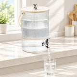 A ribbed clear glass water dispenser with a light wood lid and metal spout, shown on a kitchen counter with water flowing into a glass cup.