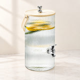 A ribbed clear glass beverage dispenser with a light wood lid, a small metal bear topper, a side handle, and a chrome spigot, shown on a light marble surface with lemon slices and water inside.