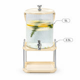 A 5L ribbed clear glass beverage dispenser with a cream-colored plastic lid and base, supported by a matching cream-colored metal stand with a lower tray. The dispenser features a chrome-finished spigot and is shown filled with water, lemon slices, and a green sprig.