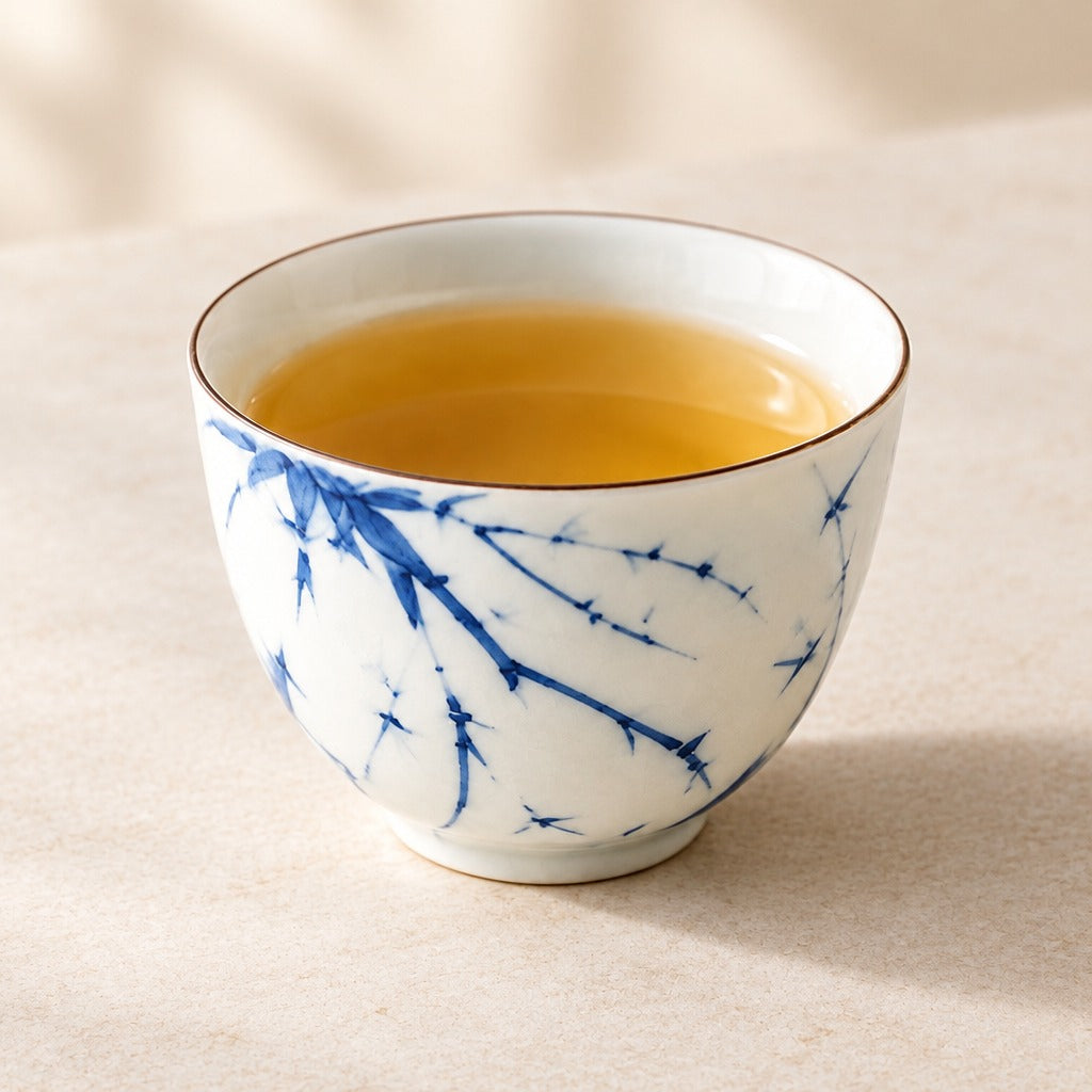A white ceramic tea cup with blue floral branch patterns and a thin gold rim, filled with amber tea, set on a beige surface with a soft-focus background.