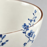 Close-up of a white ceramic bowl featuring a delicate blue floral branch pattern and a thin brown rim, shown against a neutral light gray background.
