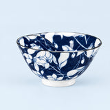 A ceramic bowl with a dark blue and white floral pattern, featuring a white footed base and a thin dark rim, set against a light blue background.