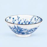A white ceramic bowl with a blue hand-painted landscape design of sailing ships on the exterior and interior, finished with a thin gold rim, set against a solid light blue background.