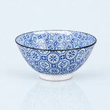 A ceramic bowl with a blue and white geometric pattern, featuring a thin black rim and a white footed base, set against a light blue background with a soft reflection.