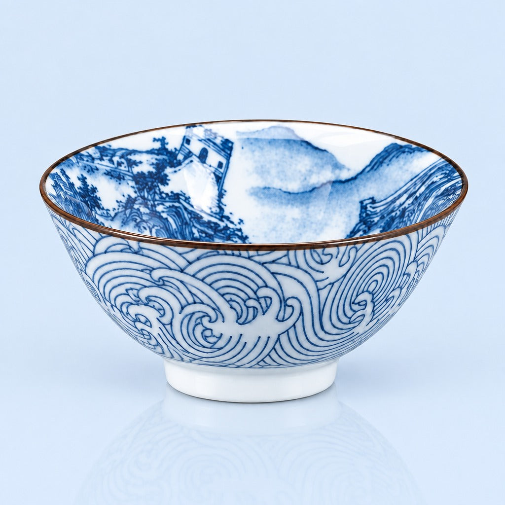 A white ceramic bowl decorated with blue hand-painted wave patterns on the exterior and a scenic landscape with a tower on the interior, finished with a thin brown rim.