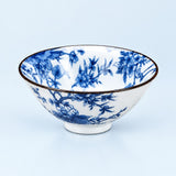 A white ceramic bowl decorated with a blue floral and bird pattern, featuring a thin dark rim and a small footed base, set against a light blue background.