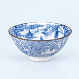A white ceramic bowl decorated with blue hand-painted traditional Chinese landscape and wave patterns, featuring a thin gold rim, set against a light blue background.