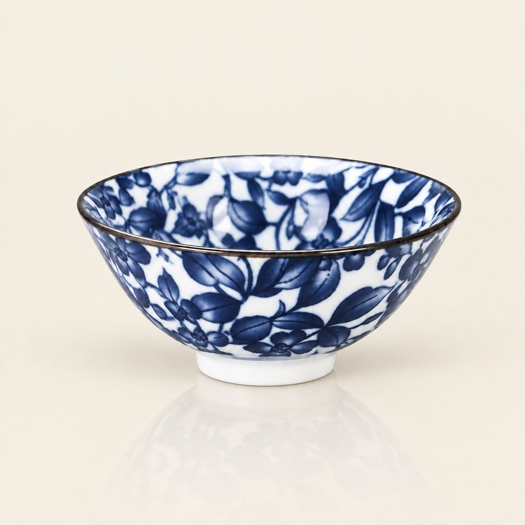 A white ceramic bowl decorated with a blue floral and leaf pattern, featuring a thin dark rim and a small white footed base, set against a warm off-white background.