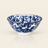 A white ceramic bowl decorated with a blue floral and leaf pattern, featuring a thin dark rim and a small white footed base, set against a warm off-white background.