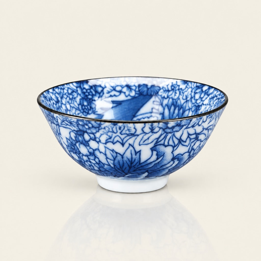 A blue and white porcelain bowl with a detailed floral pattern and a thin black rim, shown on a light beige background with a soft reflection below.